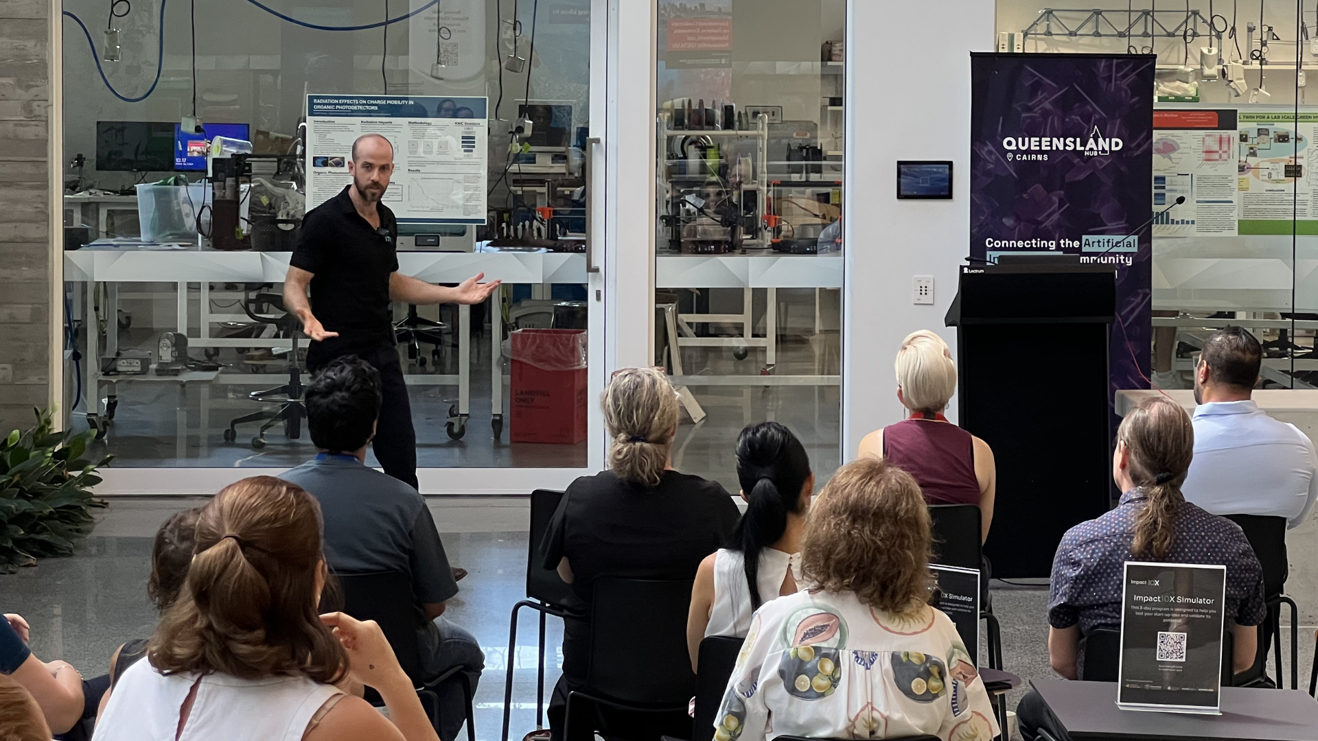 Man in a black polo presenting to a seated audience in a modern lab, with lab benches and equipment visible behind a glass wall.