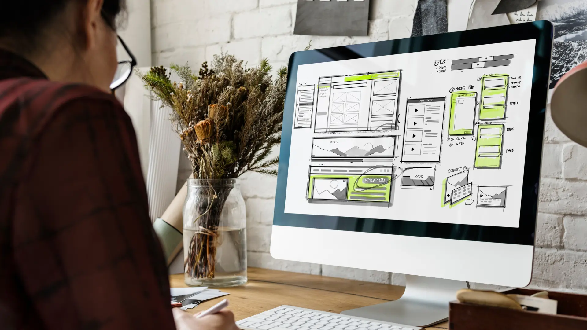 Person sketching a website wireframe on a large desktop monitor in a bright workspace, with a vase of dried plants nearby.