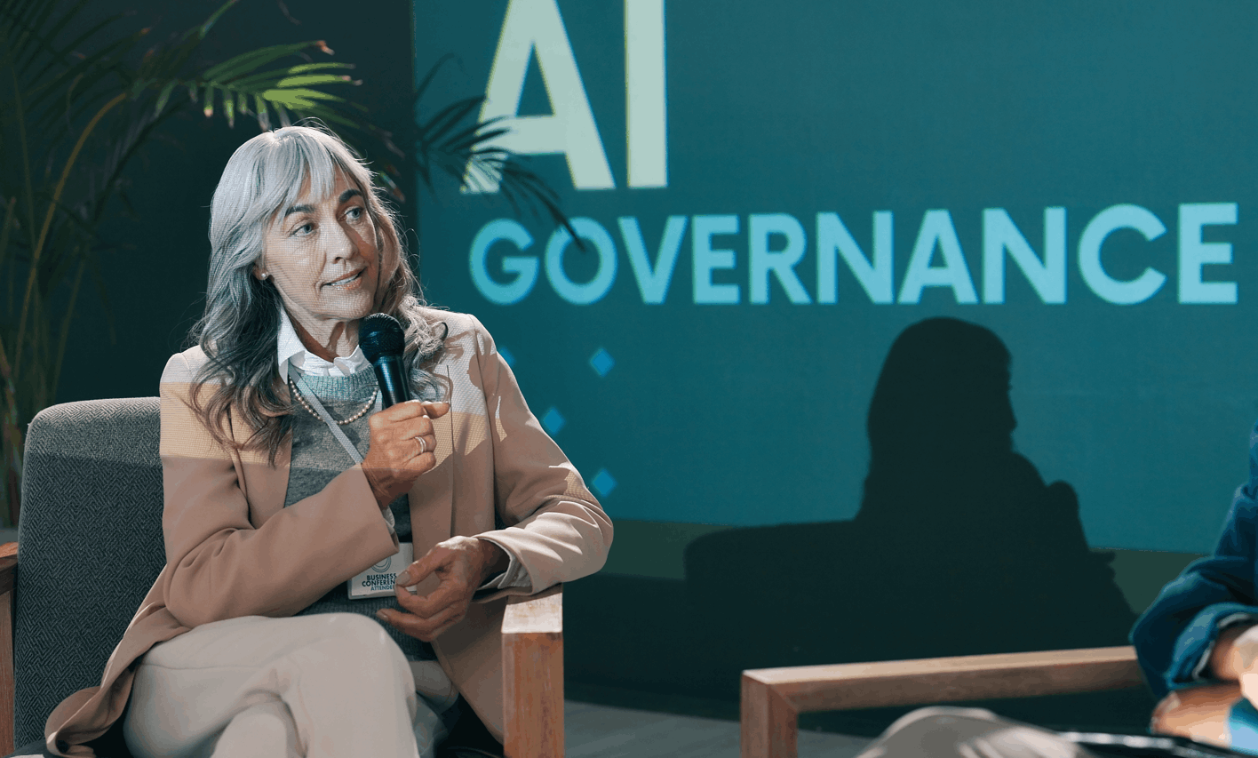 Seated woman with gray hair speaks into a handheld microphone during a panel discussion, badge visible.