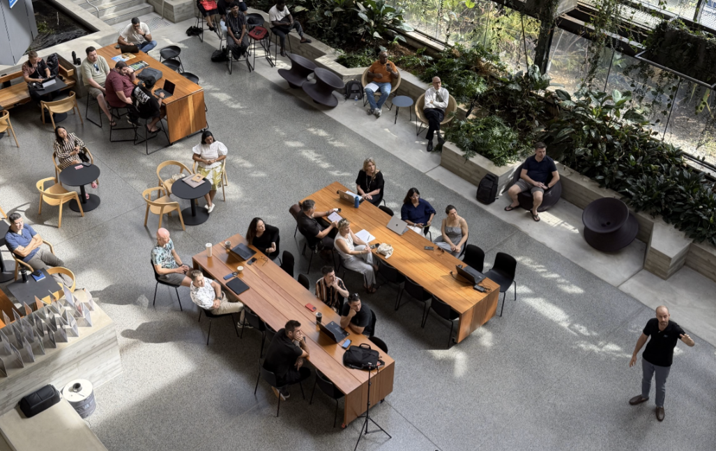 Top-down view of a coworking space with groups of people at long wooden tables using laptops; a presenter stands on the right side.